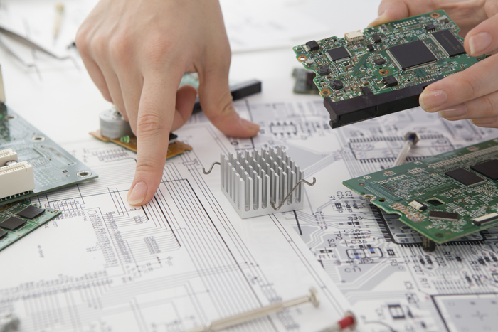 Hand zeigt auf Schaltplan, umgeben von Leiterplatten und Kühlkörpern – technische Entwicklung von Embedded Systemen